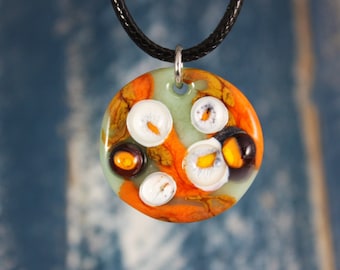 May include: A round glass pendant necklace with a green, orange, and white abstract design. The pendant has a silver bail and is hanging from a black cord.