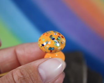 Può includere: Un piccolo perno di vetro di forma a fungo, di colore arancione, macchiato con un design a coriandoli bianco e multicolore.