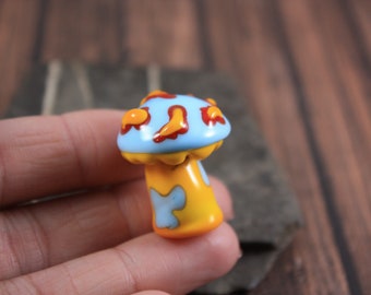 Può includere: Un piccolo fungo di vetro dipinto a mano con un cappello blu e un gambo giallo. Il cappello ha accenti rossi e arancioni.