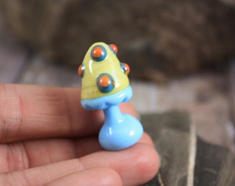 Può includere: Un piccolo perno di vetro a forma di fungo con un cappello giallo e uno stelo blu. Il cappello ha pois arancioni e blu.