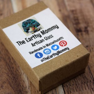 May include: A brown cardboard box with a white label that says "The Earthy Mommy Artisan Glass Handmade in Massachusetts" and includes social media icons for Facebook, Instagram, Twitter, and Pinterest. The text "@TheEarthyMommy" is also on the label.