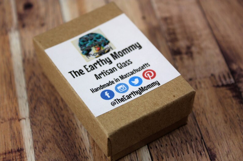 May include: A brown cardboard box with a white label that says "The Earthy Mommy Artisan Glass Handmade in Massachusetts" and includes social media icons for Facebook, Instagram, Twitter, and Pinterest. The text "@TheEarthyMommy" is also on the label.