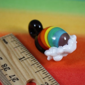 May include: A glass rainbow charm with a white cloud design. The charm is made of glass and has a rainbow pattern with a brown center. The charm is about 1 inch in diameter.