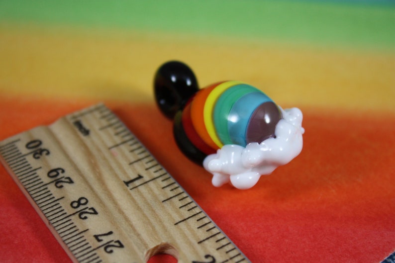 May include: A glass rainbow charm with a white cloud design. The charm is made of glass and has a rainbow pattern with a brown center. The charm is about 1 inch in diameter.