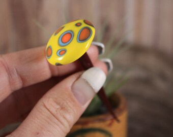 Può includere: Uno spillo in ceramica giallo con un gambo marrone. La parte superiore dello spillo è decorata con cerchi blu e arancioni.
