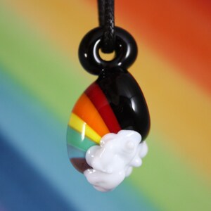 May include: A glass pendant with a rainbow design and a white cloud. The pendant is attached to a black cord.