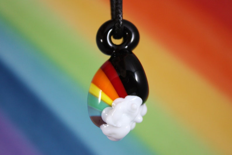 May include: A glass pendant with a rainbow design and a white cloud. The pendant is attached to a black cord.