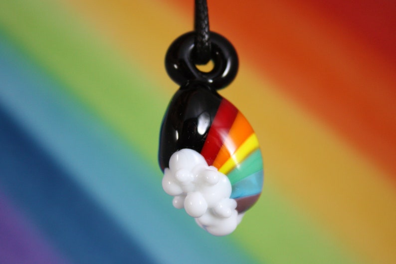 May include: A glass pendant with a rainbow design and a white cloud. The pendant is black on the top and has a black cord.