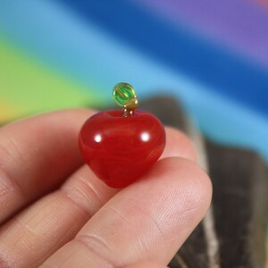 May include: A small, red glass apple with a green glass stem. The apple is being held in a hand against a blurred rainbow background.