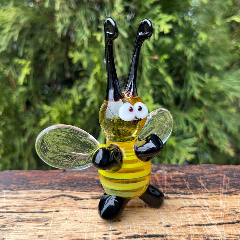 Puede incluir: Figura de abeja de cristal hecha a mano con cuerpo a rayas amarillas y negras, alas transparentes y antenas y patas negras. La abeja tiene ojos grandes y expresivos y est&aacute; posada como si estuviera de pie. El fondo es un follaje verde borroso.
