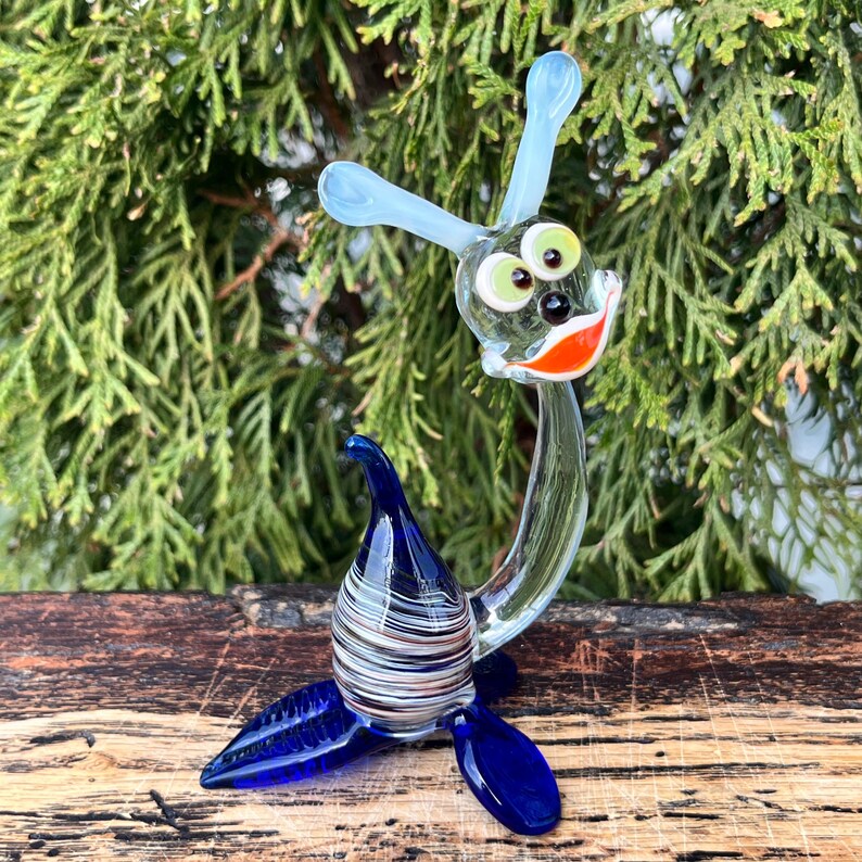 May include: A whimsical glass sculpture of a sea creature with a smiling face, blue antennae, and fins. The body is a mix of blue and white stripes. The eyes are green with black pupils, and the mouth is orange.