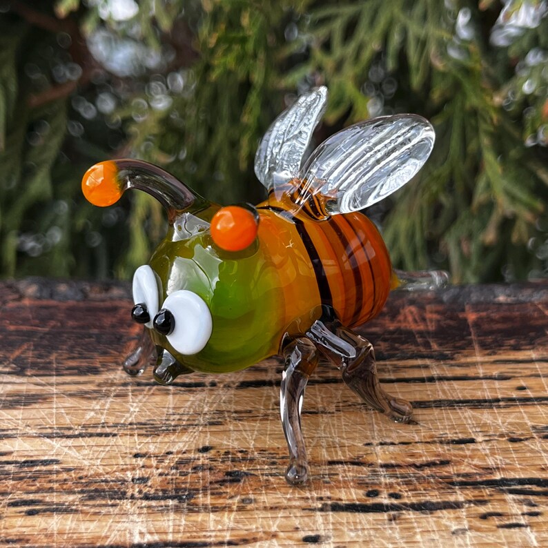 Puede incluir: Figura de abeja de vidrio hecha a mano con colores naranja, verde y &aacute;mbar. La abeja tiene alas transparentes, ojos naranjas y rayas negras y &aacute;mbar en el abdomen. La figura est&aacute; colocada sobre una superficie de madera.