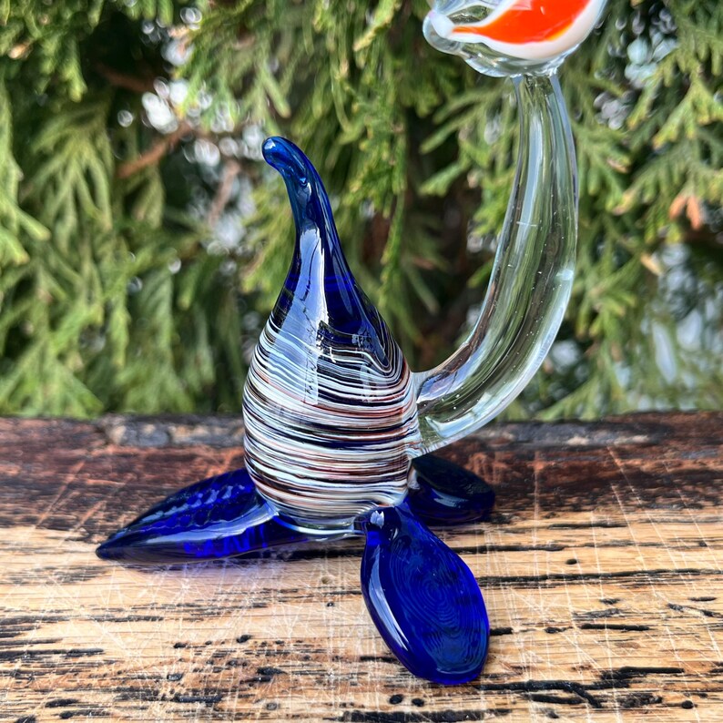 May include: A handcrafted glass sculpture of a bird with a long neck and tail. The bird features a blue head and tail, a body with white, brown, and clear stripes, and an orange and white head. The sculpture is displayed on a wooden surface.