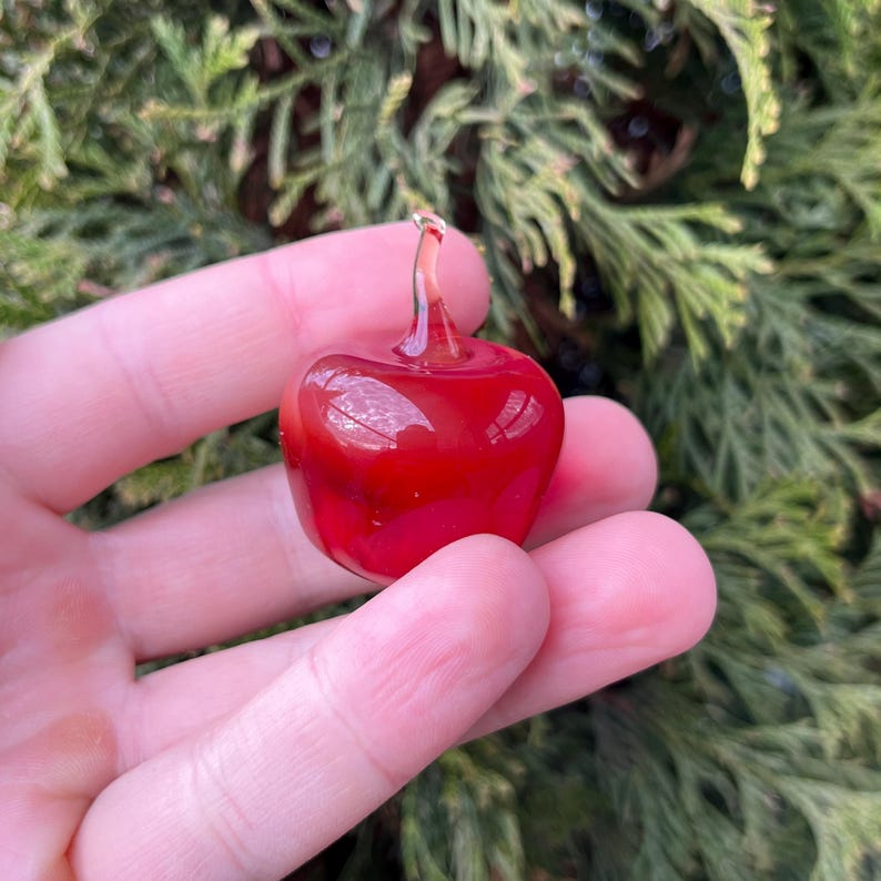 May include: A small, glossy, bright red glass apple figurine with a smooth, rounded body and a clear, curved glass stem extending from the top. The surface of the glass reflects light, showing a polished finish and a vibrant, solid red color.