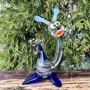 May include: A whimsical glass sculpture of a creature with a blue body, clear neck, and a head with large green eyes and an orange mouth. The sculpture has blue antennae and is set against a backdrop of green foliage.
