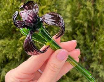 Könnte beinhalten: Eine Hand hält eine Glasblume mit einer violetten Blüte und einem grünen Stiel. Die Blume ist aus Glas und hat ein zartes, filigranes Design.