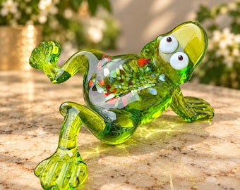 May include: A translucent green glass frog figurine featuring large white eyes with dark pupils and a body decorated with multicolored flecks of red, blue, and yellow glass embedded within the clear green structure, posed in a reclining position.
