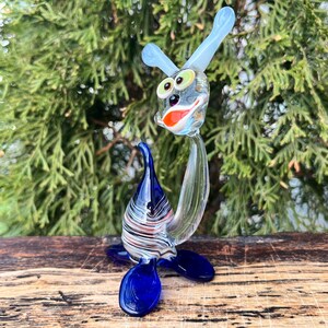 May include: A whimsical glass sculpture of a sea creature with a blue body and fins, a clear body, and a head with large green eyes, blue ears, and a red and white mouth. The sculpture is set against a backdrop of green foliage.