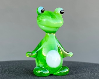May include: A small green glass frog figurine with white eyes and a white belly. The frog is standing on a black surface.