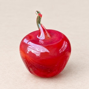 May include: A small, round, hand-blown glass apple figurine in a vibrant, translucent red color. The apple features a smooth, glossy surface with subtle internal swirls of deeper red and a clear, curved glass stem extending from the top center.