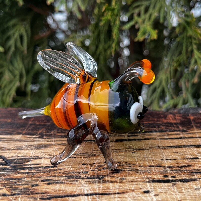Puede incluir: Figura de abeja de cristal hecha a mano con alas transparentes, cuerpo a rayas naranja y amarillo y ojos negros y blancos. La abeja tiene patas de cristal transparente y est&aacute; sobre una superficie de madera.