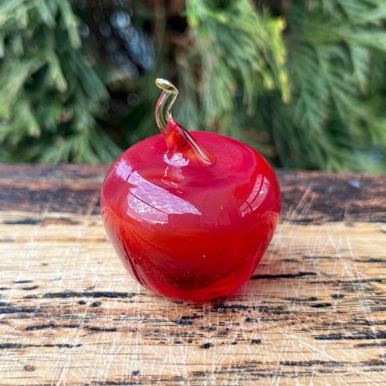 May include: A small, smooth, hand-blown glass apple figurine in a vibrant, translucent red color. The apple features a clear, curved glass stem extending from the top center, showcasing a glossy, reflective surface and rounded shape.