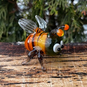 Puede incluir: Figurita de abeja de cristal hecha a mano con alas transparentes, antenas naranjas y cuerpo a rayas en tonos naranja y amarillo. La abeja tiene ojos blancos y patas oscuras, y se exhibe sobre una superficie de madera.