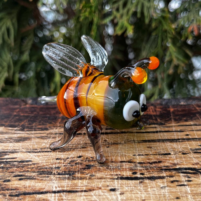 Puede incluir: Figurita de abeja de cristal hecha a mano con alas transparentes, antenas naranjas y cuerpo a rayas en tonos naranja y amarillo. La abeja tiene ojos blancos y patas oscuras, y se exhibe sobre una superficie de madera.