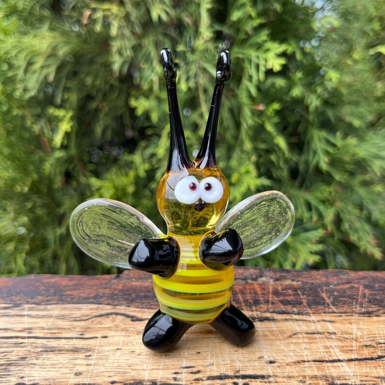 Puede incluir: Una figura de abeja de vidrio hecha a mano con rayas amarillas y negras, alas transparentes y antenas negras. La abeja tiene grandes ojos blancos y patas negras. La figura se exhibe sobre una superficie de madera con un fondo verde.