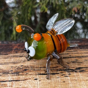 Puede incluir: Figura de abeja de cristal colorida, con tonos naranja, verde y amarillo. La abeja tiene alas transparentes, ojos naranjas y rayas negras en el abdomen. Posada sobre una superficie de madera.
