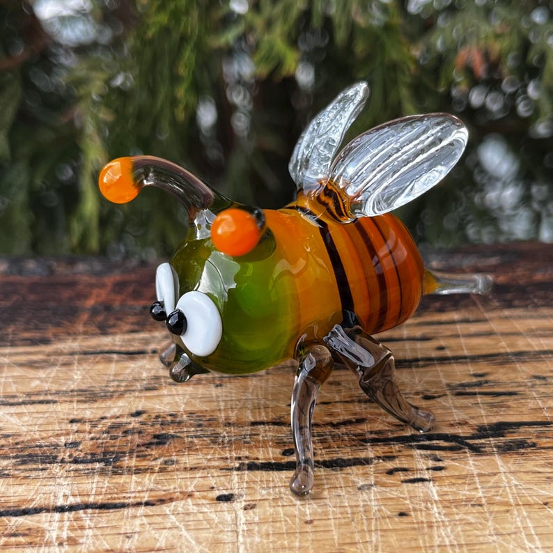Puede incluir: Figura de abeja de cristal colorida, con tonos naranja, verde y amarillo. La abeja tiene alas transparentes, ojos naranjas y rayas negras en el abdomen. Posada sobre una superficie de madera.