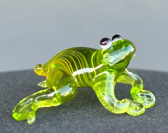 May include: A small, green glass frog figurine with black eyes. The frog has a striped pattern and is sitting on a dark surface.