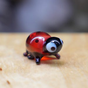May include: A small, glass ladybug figurine. The ladybug has a red body with black spots and a black head with white and brown eyes. The legs are black. The figurine is sitting on a wooden surface.