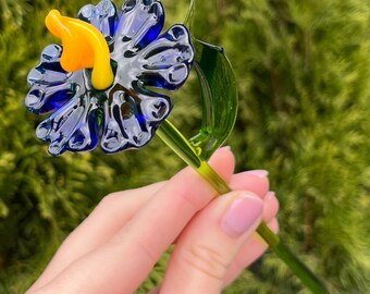 May include: A blue glass flower with a yellow center and green stem. The flower is held in a hand.