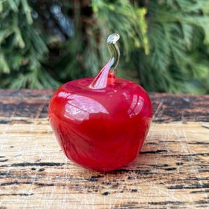 May include: A small, solid glass apple figurine in a vibrant, translucent red hue. The apple features a smooth, rounded body and a clear, curved glass stem extending from the top, showcasing a glossy finish and reflective surface quality.