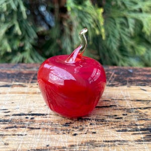 May include: A small, smooth, bright red glass apple figurine with a clear, curved glass stem extending from the top. The object has a glossy, reflective surface and a rounded, bulbous shape, capturing light across its vibrant red body.