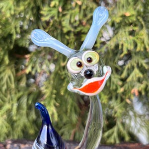 May include: A whimsical glass sculpture of a smiling creature with large eyes, a black nose, and an orange and white mouth. The sculpture has light blue antennae and a dark blue and white striped base. The background is a blurred view of green foliage.