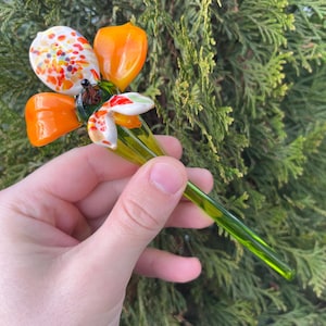 Könnte beinhalten: Eine handgefertigte Glasblume mit orangefarbenen, weißen und grünen Farbtönen. Die Blume hat eine gesprenkelte Mitte und orangefarbene Blütenblätter. Der Stiel ist ein leuchtendes Grün. Die Blume wird vor einem Hintergrund aus grünem Laub gehalten.