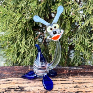 May include: A whimsical glass sculpture of a cartoon-like creature with a blue head, ears, and tail. The sculpture has a clear body, a red and white striped base, and a smiling face with green eyes and an orange mouth. The sculpture is on a wooden surface.
