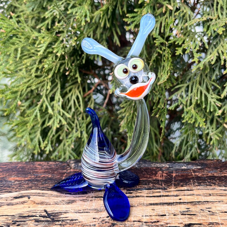 May include: A whimsical glass sculpture of a cartoon-like creature with a blue head, ears, and tail. The sculpture has a clear body, a red and white striped base, and a smiling face with green eyes and an orange mouth. The sculpture is on a wooden surface.
