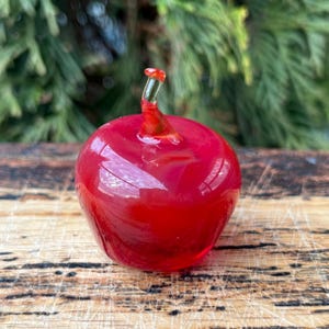 May include: A small, solid glass apple figurine in a vibrant, translucent red hue. The apple features a smooth, rounded body and a clear, curved glass stem extending from the top center, reflecting light across its polished, glossy surface.