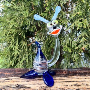 May include: A whimsical glass sculpture of a sea creature with a smiling face, blue antennae, and flipper-like feet. The body is clear with a blue, white, and red striped design. The sculpture is set against a backdrop of green foliage.