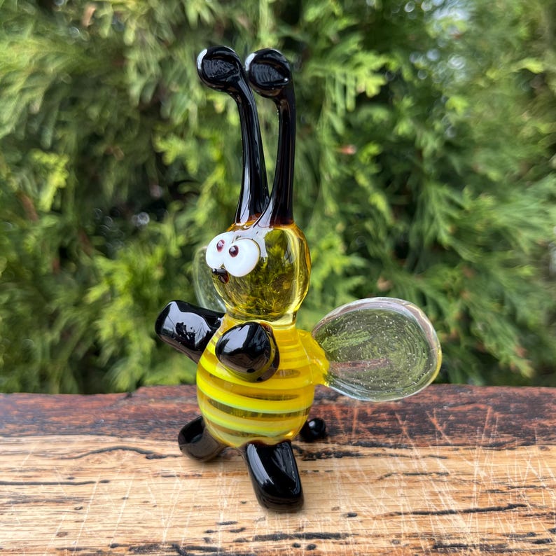 Puede incluir: Figura de abeja de cristal hecha a mano. La abeja tiene un cuerpo a rayas amarillas y negras, patas y antenas negras y alas transparentes. Los ojos son blancos con pupilas negras. Se muestra sobre una superficie de madera con fondo verde.