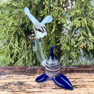 May include: A glass sculpture of a seal with a clear body and head, and blue flippers and base. The seal's body has horizontal stripes in various colors. The sculpture is displayed on a wooden surface with a green background.