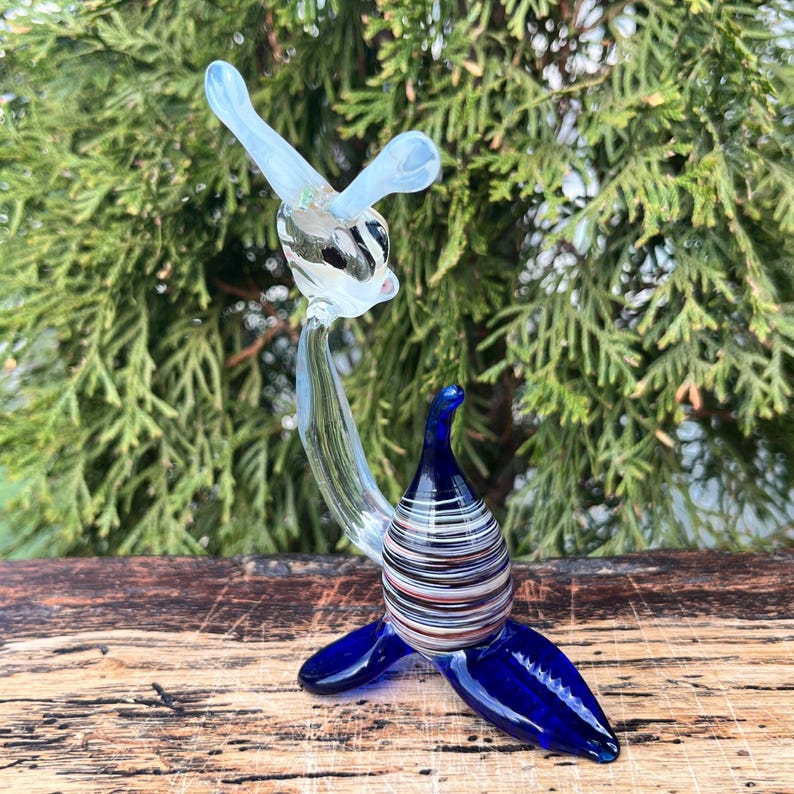 May include: A glass sculpture of a seal with a clear body and head, and blue flippers and base. The seal's body has horizontal stripes in various colors. The sculpture is displayed on a wooden surface with a green background.