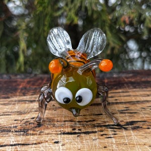 Puede incluir: Figura de abeja de cristal hecha a mano con cuerpo verde, detalles naranjas y alas transparentes. La abeja tiene ojos blancos grandes con pupilas negras y est&aacute; posada sobre una superficie de madera. El cuerpo de la abeja presenta rayas naranjas y negras.