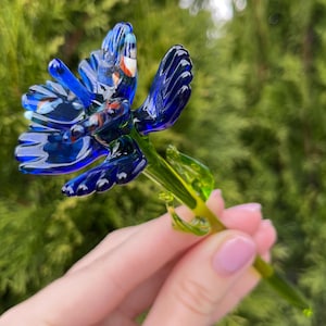 Può includere: Un fiore di vetro blu con stelo e foglie verdi. Il fiore ha un design dettagliato con accenti blu, bianchi e rossi.