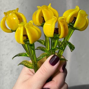 May include: A bouquet of yellow glass flowers with green stems and leaves. The flowers are in full bloom, showcasing their intricate details. The glass is transparent, allowing light to pass through, and the bouquet is held in a hand.