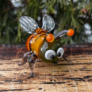 Puede incluir: Figura de abeja de cristal hecha a mano. La abeja tiene una cabeza verde, un cuerpo a rayas naranja y amarillo, alas transparentes y antenas naranjas. La figura est&aacute; sobre una superficie de madera.