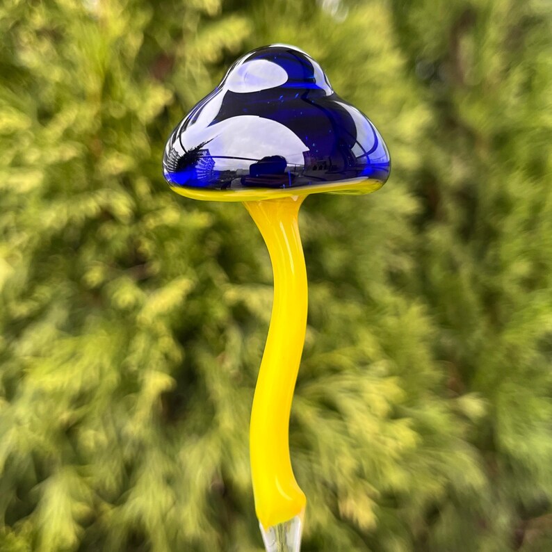 May include: A glass mushroom garden stake with a blue cap and a yellow stem. The mushroom is made of blown glass and has a unique, whimsical design.